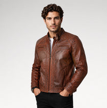 A man wearing a brown distressed leather jacket over a white t-shirt and black pants, standing against a light gray studio background.