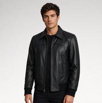 A man wearing a black leather jacket with a zippered front and collar, paired with a black t-shirt and black pants, photographed against a light gray studio background.