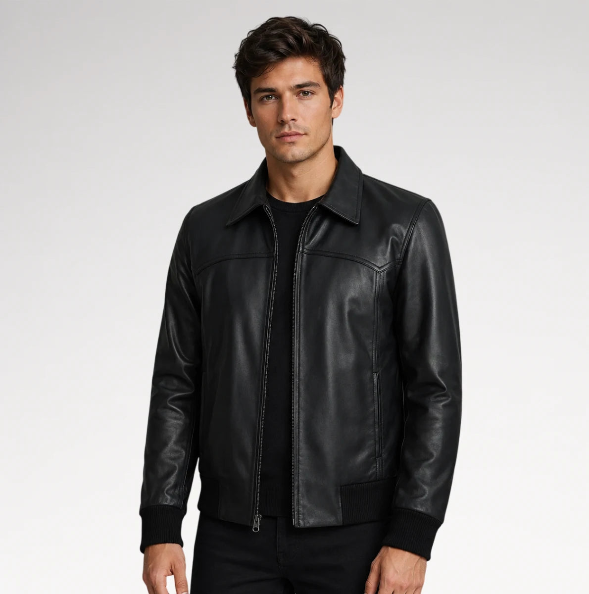 A man wearing a black leather jacket with a zippered front and collar, paired with a black t-shirt and black pants, photographed against a light gray studio background.