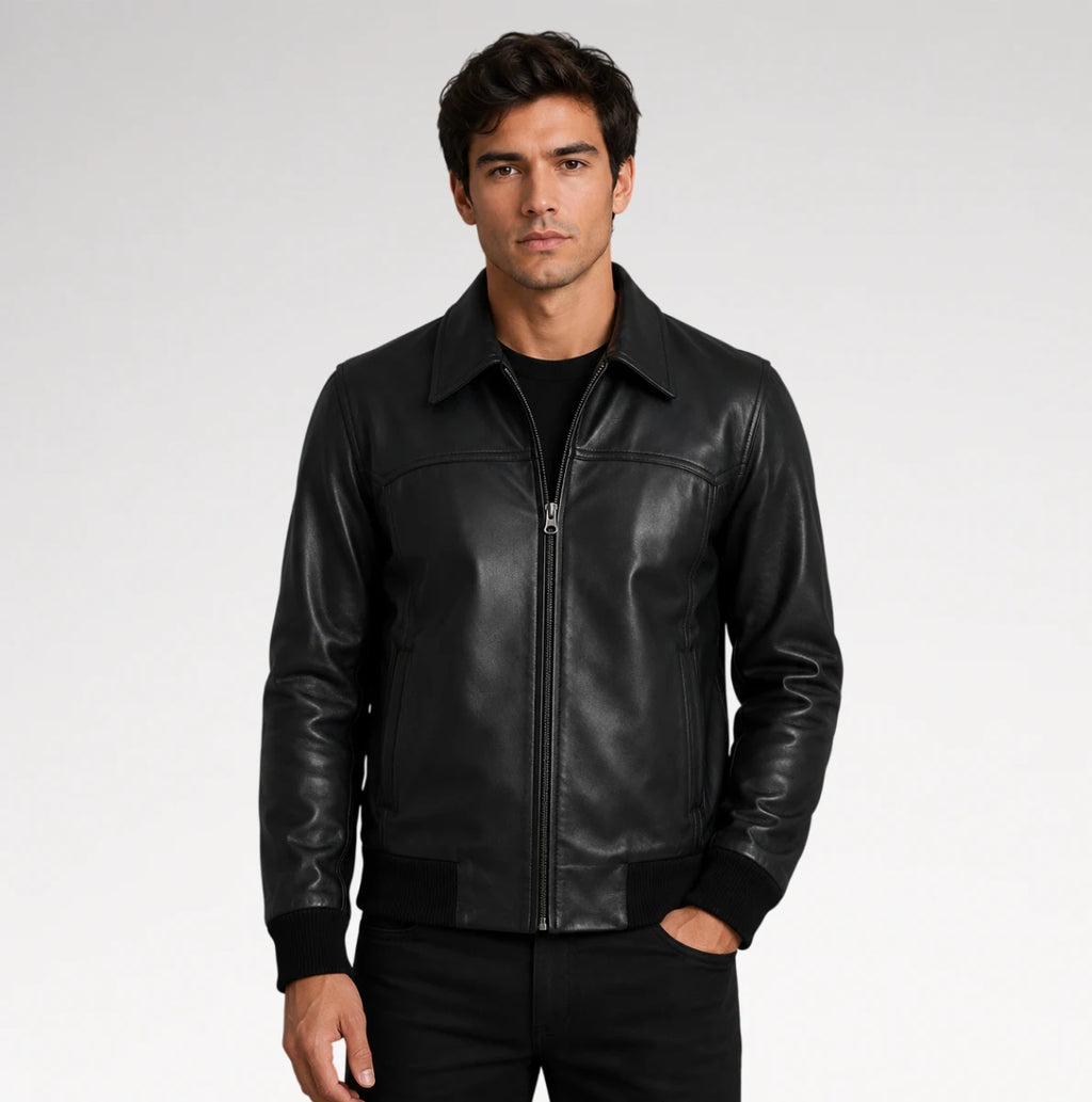 A man wearing a black leather jacket with a front zipper, ribbed collar, cuffs, and hem, paired with a black t-shirt and black pants, photographed against a light gray studio background.