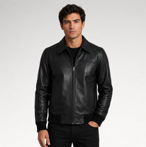 A man wearing a black leather jacket with a front zipper, ribbed collar, cuffs, and hem, paired with a black t-shirt and black pants, photographed against a light gray studio background.