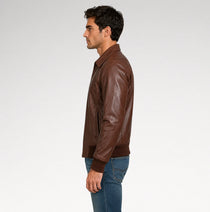 A man in profile wearing a brown leather jacket and blue jeans, photographed against a plain light gray studio background.