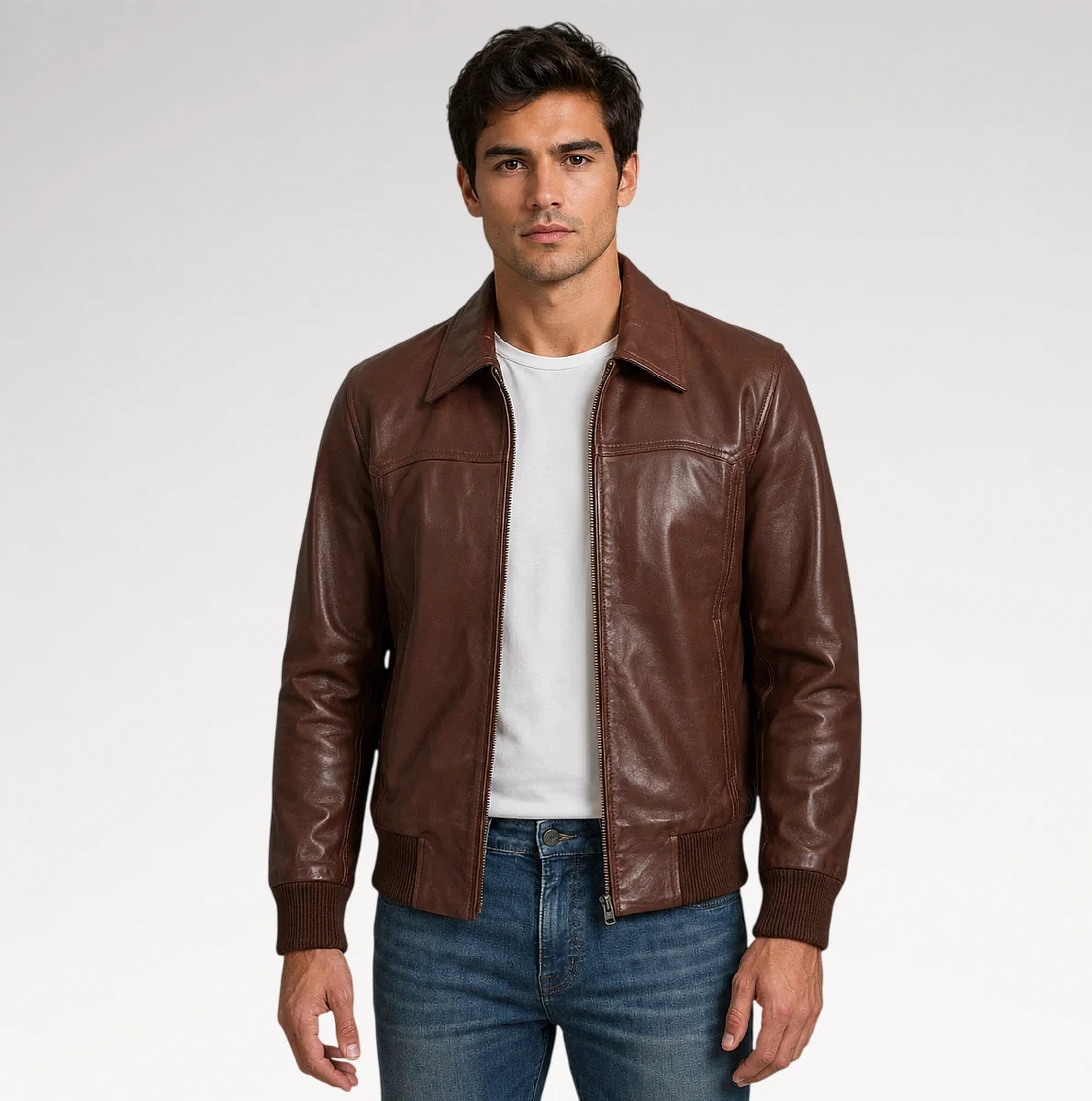 A man wearing a brown leather jacket over a white t-shirt and blue jeans, standing against a light gray studio background. Main image