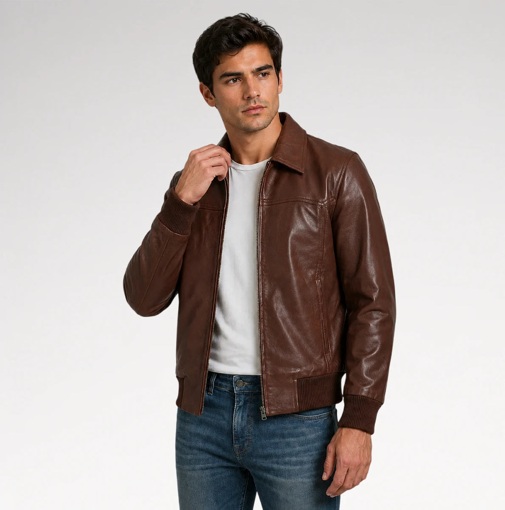A man wearing a brown leather jacket over a white t-shirt and blue jeans, standing against a light gray studio background.