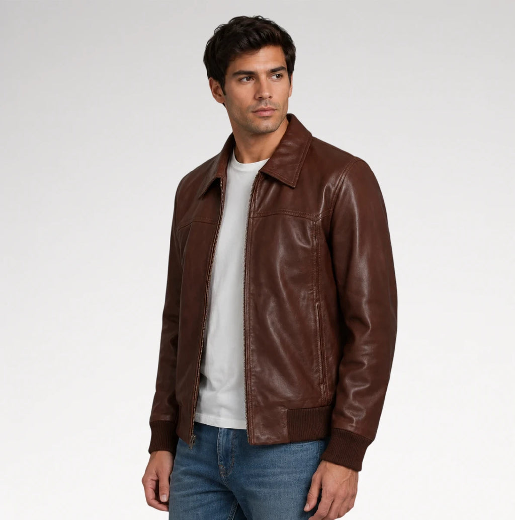 A man wearing a brown leather jacket over a white t-shirt and blue jeans, standing against a light gray studio background.