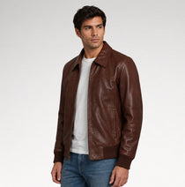 A man wearing a brown leather jacket over a white t-shirt and blue jeans, standing against a light gray studio background.