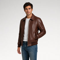 A man wearing a brown leather jacket over a white t-shirt and blue jeans, standing against a light gray studio background.