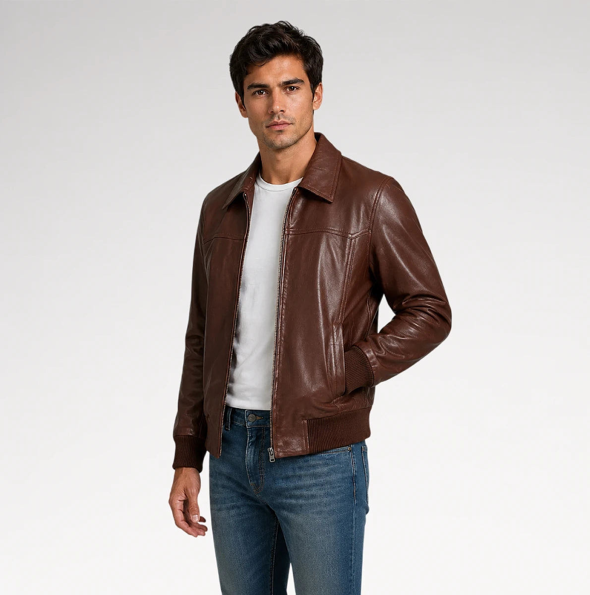A man wearing a brown leather jacket over a white t-shirt and blue jeans, standing against a light gray studio background.