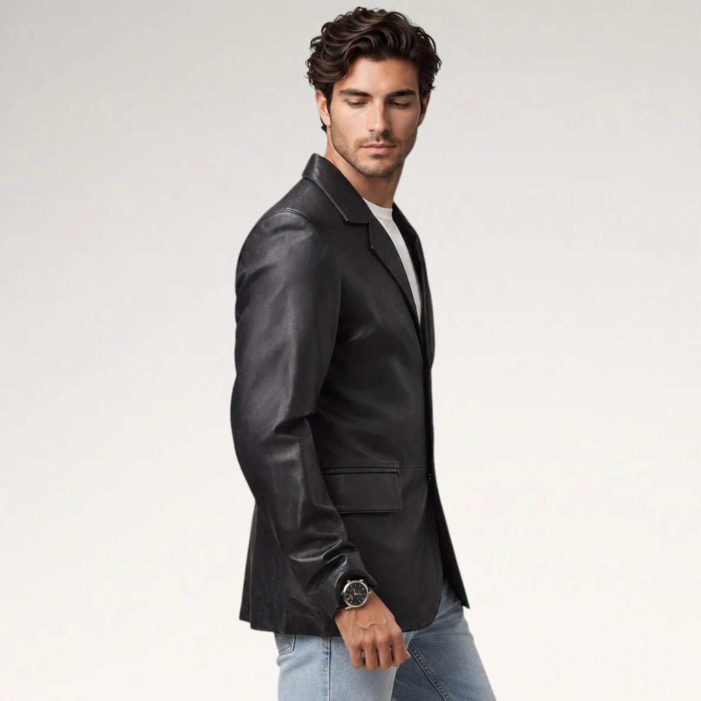 A man wearing a black leather blazer over a white t-shirt and light blue jeans, standing against a light gray studio background.