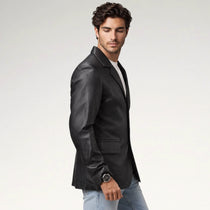 A man wearing a black leather blazer over a white t-shirt and light blue jeans, standing against a light gray studio background.