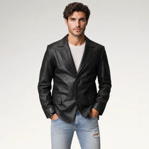 A man wearing a black leather blazer over a white t-shirt and light blue jeans, standing against a light gray studio background.