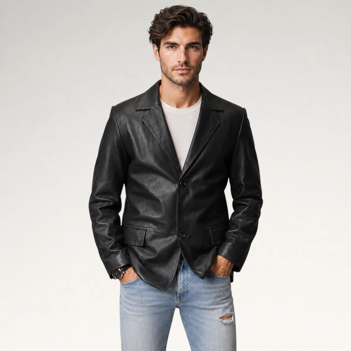 A man wearing a black leather blazer over a white t-shirt and light blue jeans, standing against a light gray studio background.