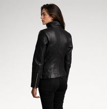 A woman shown from the back wearing a black leather jacket with quilted sleeves and a high collar, paired with black pants, against a plain light gray studio background.