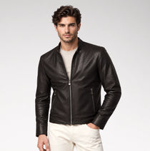 A man wearing a black leather moto jacket over a light beige t-shirt and off-white trousers, standing against a light gray studio background.