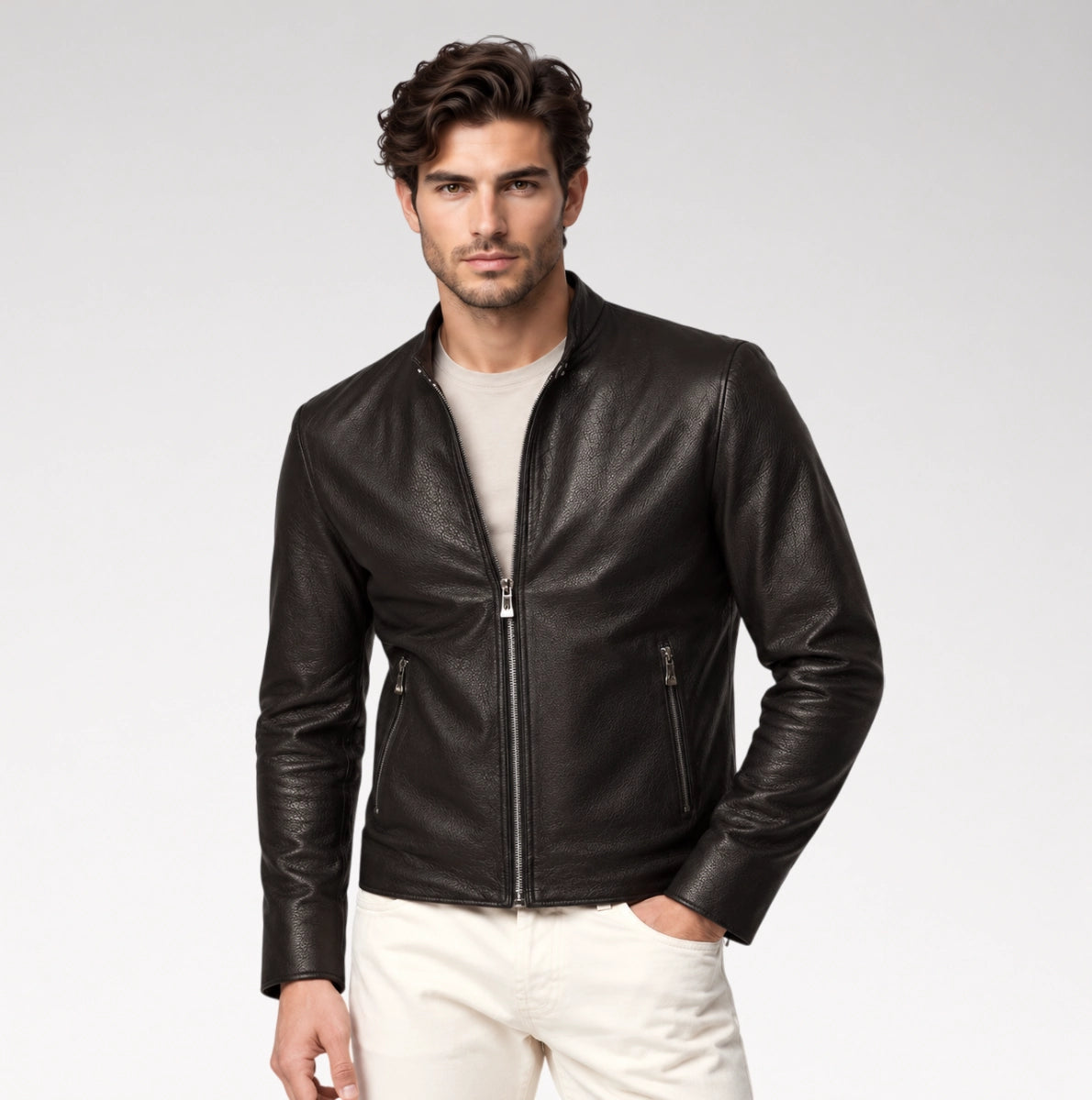 A man wearing a black leather moto jacket over a light beige t-shirt and off-white trousers, standing against a light gray studio background.