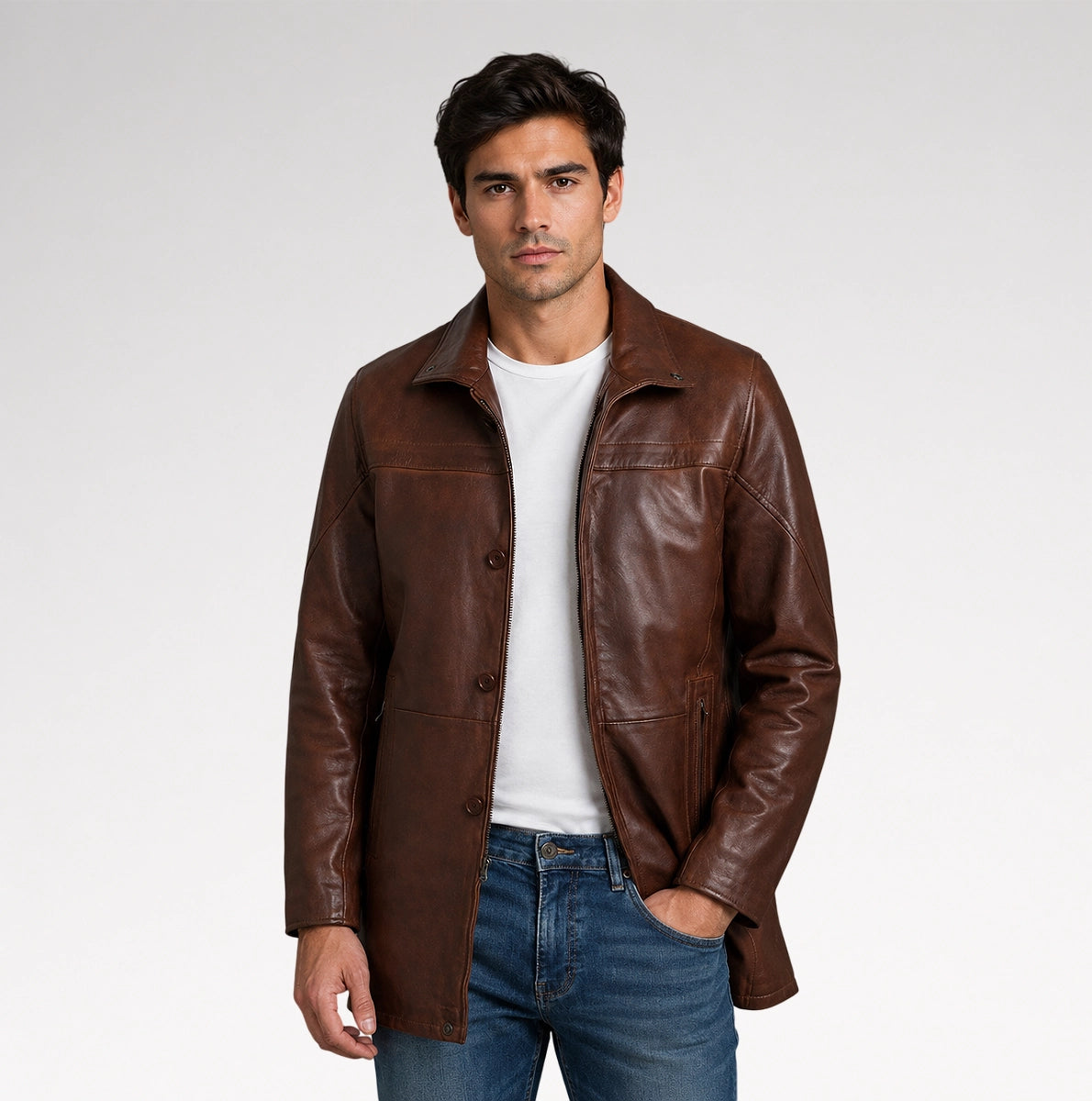 A man wearing a brown leather jacket over a white t-shirt and blue jeans, standing against a light gray studio background.