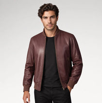 A man wearing a dark brown leather bomber jacket over a black t-shirt and black pants, standing against a light gray studio background.