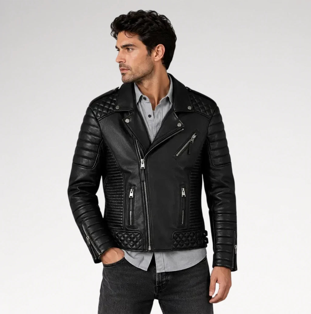 A man wearing a black leather biker jacket with quilted shoulder panels, ribbed sleeves, and silver zippers, styled over a light gray button-down shirt and dark jeans against a light gray studio background.