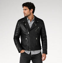 A man wearing a black leather biker jacket with quilted shoulder panels, ribbed sleeves, and silver zippers, styled over a light gray button-down shirt and dark jeans against a light gray studio background.