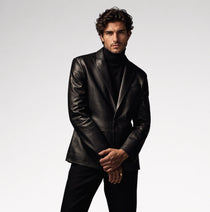 A man wearing a black leather blazer over a black turtleneck sweater and black trousers, standing against a light gray studio background.