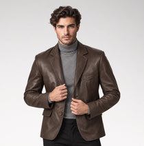 A man wearing a brown leather blazer over a gray turtleneck sweater and black trousers, standing against a light gray studio background.