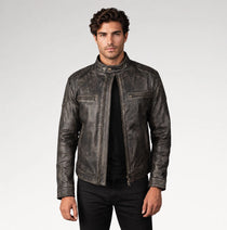 A man wearing a distressed black leather motorcycle jacket over a black shirt and black pants, standing against a light gray studio background.