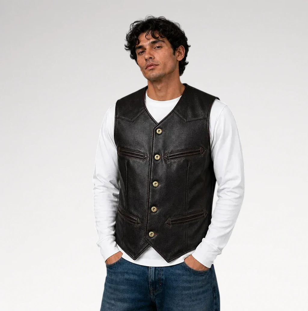 A man wearing a black leather vest over a white long-sleeve shirt and blue jeans, standing against a light gray studio background.