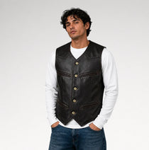 A man wearing a black leather vest over a white long-sleeve shirt and blue jeans, standing against a light gray studio background.