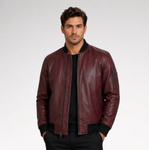 A man wearing a burgundy leather bomber jacket with black ribbed trim over a black shirt, photographed against a light gray studio background.