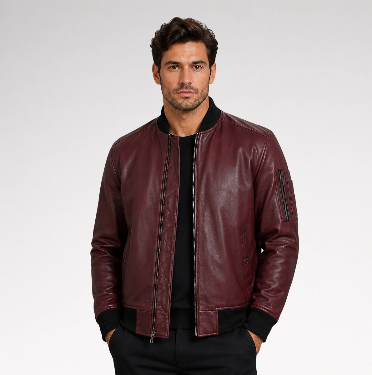 A man wearing a burgundy leather bomber jacket with black ribbed trim over a black shirt, photographed against a light gray studio background.
