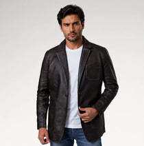 A man wearing a dark brown leather blazer over a white t-shirt and blue jeans, standing against a light gray studio background.