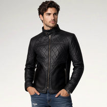 A man wearing a black quilted leather jacket with a high collar and front zipper, paired with blue jeans, photographed against a light gray studio background.