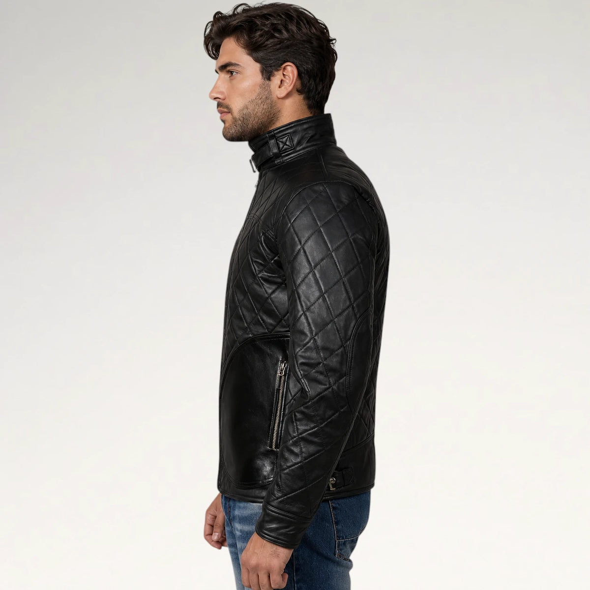 A man in profile wearing a black quilted leather jacket with zippered pockets, paired with blue jeans, against a plain light gray studio background.