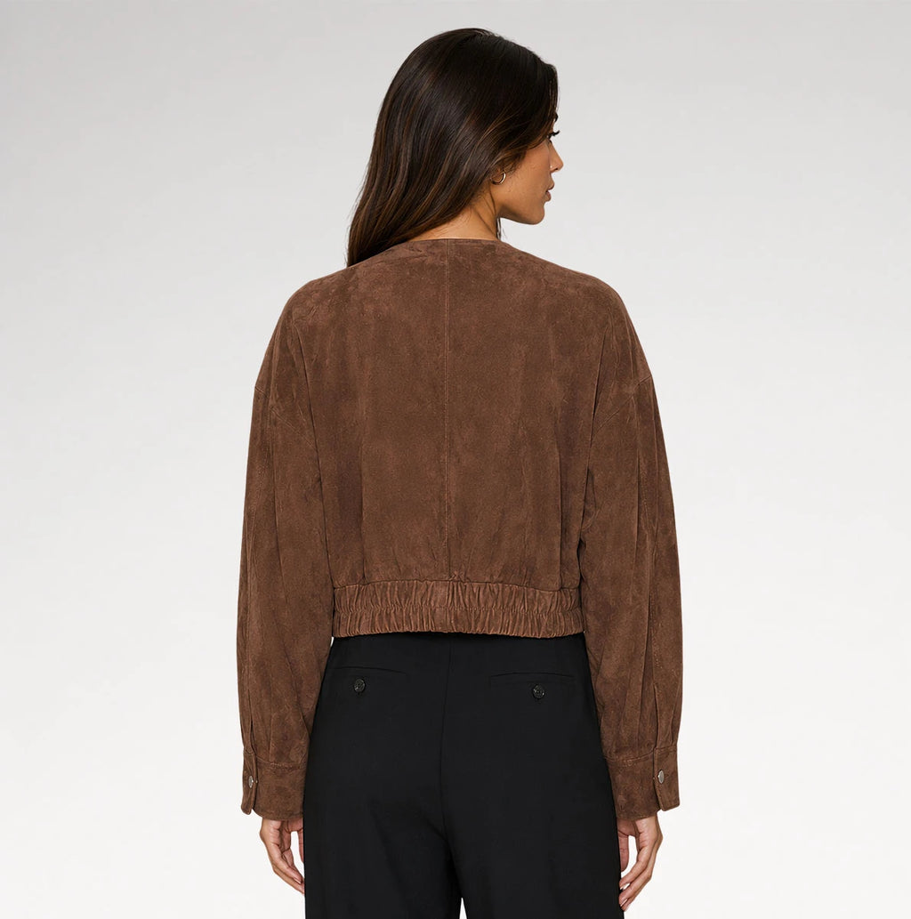 A woman shown from the back wearing a cropped brown suede jacket and black trousers against a light gray studio background.