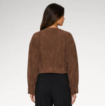 A woman shown from the back wearing a cropped brown suede jacket and black trousers against a light gray studio background.