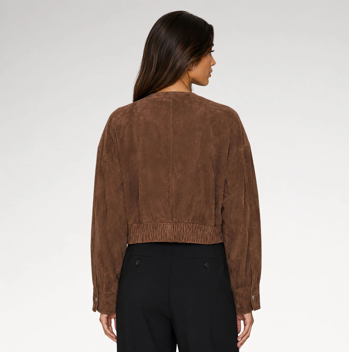 A woman shown from the back wearing a cropped brown suede jacket and black trousers against a light gray studio background.
