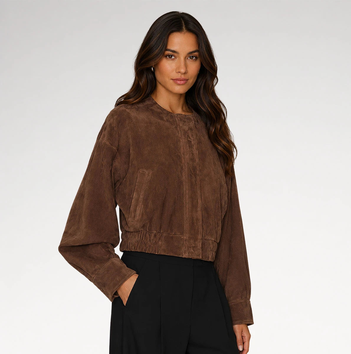 A woman modeling a cropped brown suede jacket with long balloon sleeves and front patch pockets, paired with black pants, against a light gray studio background.