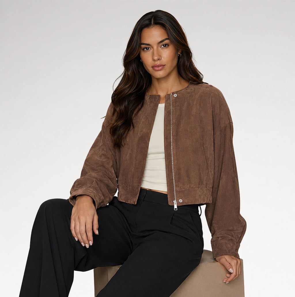 A woman models a brown cropped suede bomber jacket over a white top and black pants, seated against a plain light gray studio background.