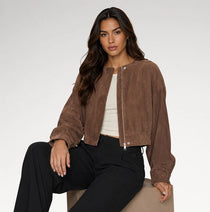 A woman models a brown cropped suede bomber jacket over a white top and black pants, seated against a plain light gray studio background.