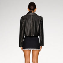 A model shown from the back wearing a black leather cropped jacket and a dark pinstriped mini skirt with a lace hem, against a light gray studio background.