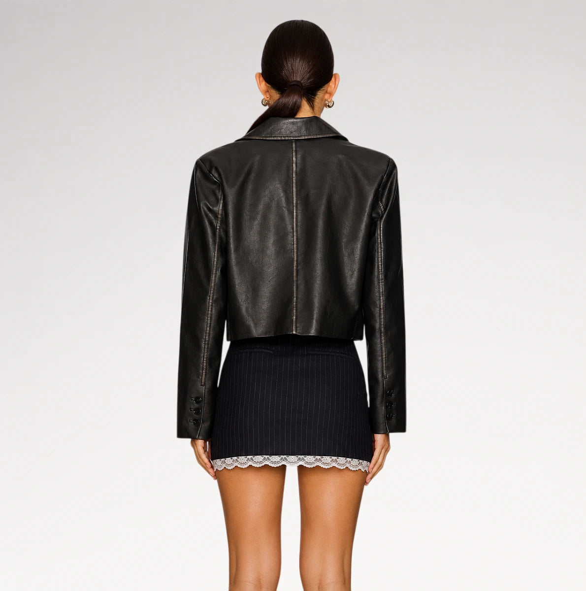 A model shown from the back wearing a black leather cropped jacket and a dark pinstriped mini skirt with a lace hem, against a light gray studio background.
