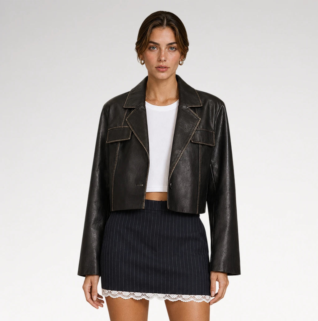 A model wearing a cropped black faux-leather jacket with contrast stitching over a white crop top and a navy blue pinstriped mini skirt with a lace hem, shot against a light gray studio background.