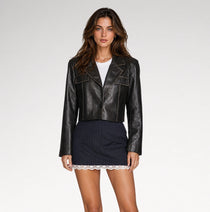 A woman modeling a black cropped leather jacket over a white top and a navy pinstriped mini skirt with a lace hem, photographed against a light gray studio background.