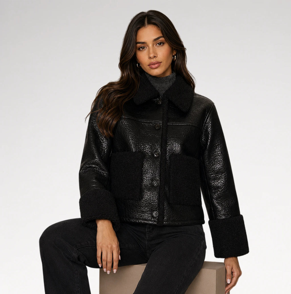 A woman models a black textured faux-leather jacket with a plush black shearling collar, button front, and large front patch pockets, paired with black jeans against a light gray studio background.