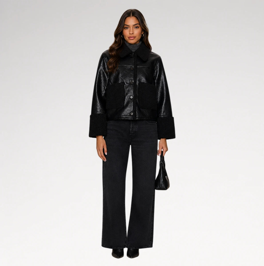 A model stands against a light gray studio background wearing a black cropped jacket with a glossy finish, wide-leg black jeans, and holding a small black handbag.