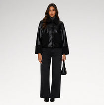 A model stands against a light gray studio background wearing a black cropped jacket with a glossy finish, wide-leg black jeans, and holding a small black handbag.