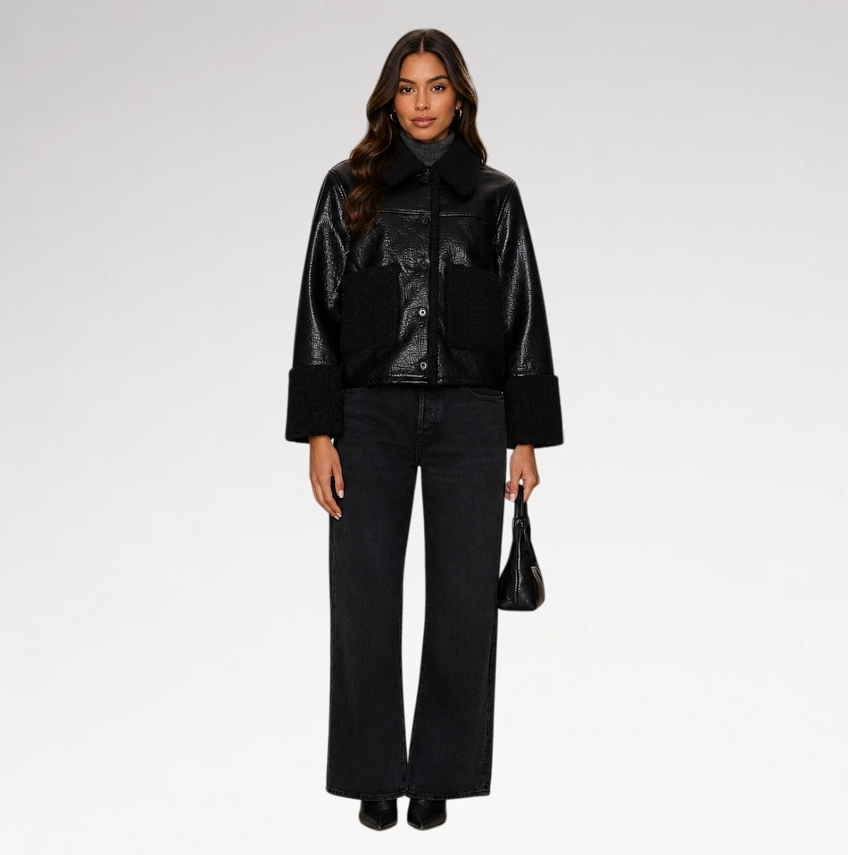 A model stands against a light gray studio background wearing a black cropped jacket with a glossy finish, wide-leg black jeans, and holding a small black handbag.