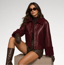 A woman models a burgundy faux-leather jacket with a textured sherpa collar and cuffs, styled with brown leather shorts, gloves, and boots against a light gray studio background.
