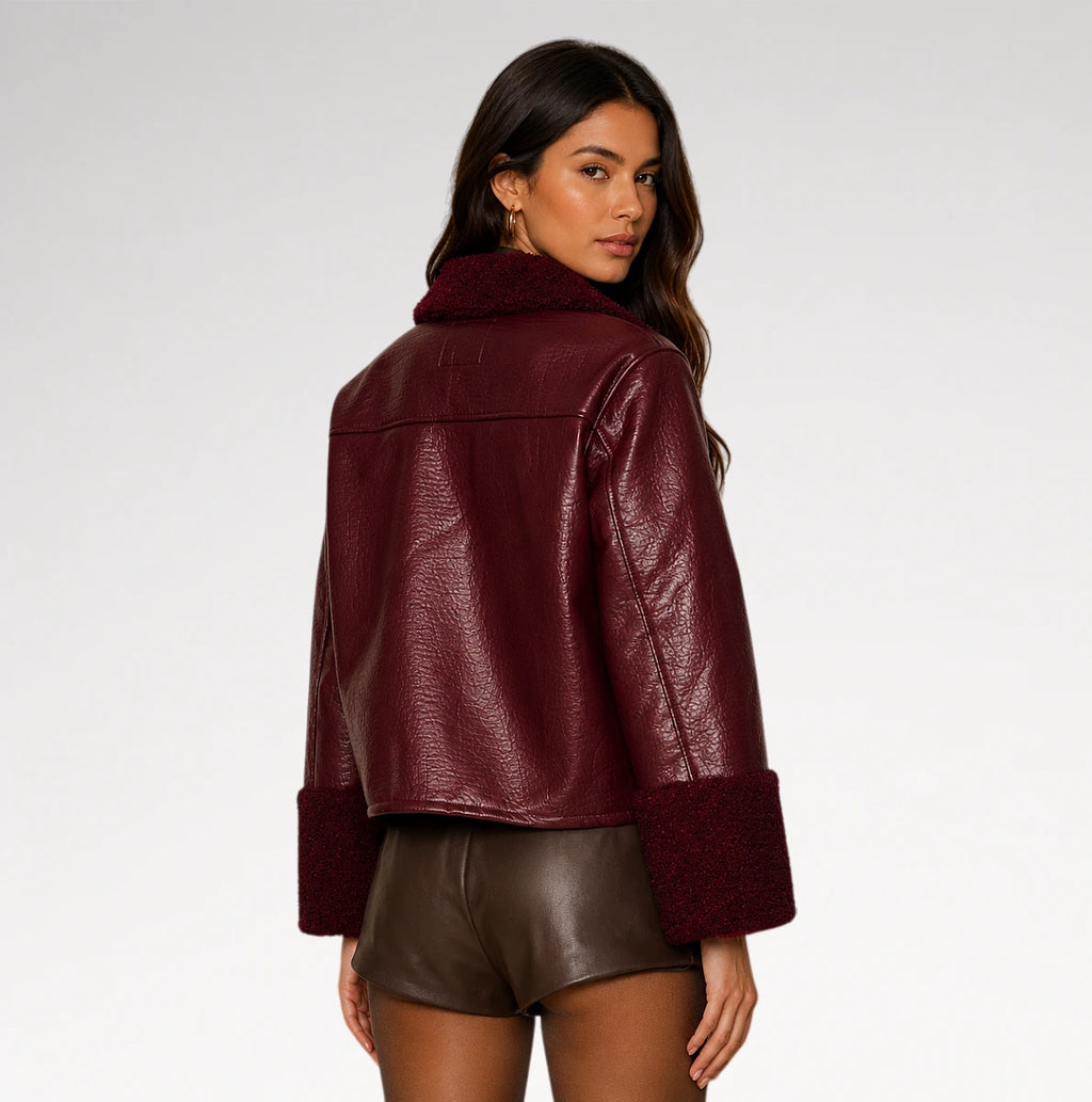 A woman shown from the back wearing a cropped, pebbled faux-leather jacket in deep burgundy with a matching faux-fur collar and cuffs, paired with brown faux-leather shorts, against a light gray studio background.