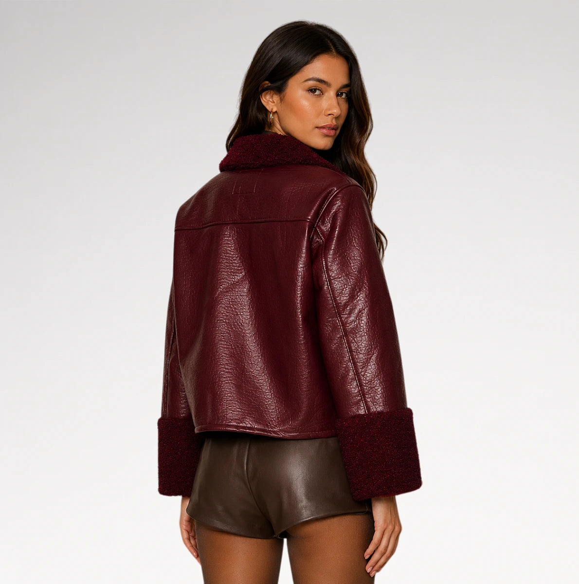 A woman shown from the back wearing a cropped, pebbled faux-leather jacket in deep burgundy with a matching faux-fur collar and cuffs, paired with brown faux-leather shorts, against a light gray studio background.